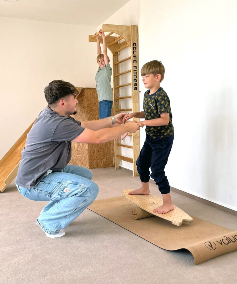 Ergotherapie Steiger Ein Mann stützt ein Kind auf einem Balanceboard, im Hintergrund turnt ein Junge an einer Sprossenwand.